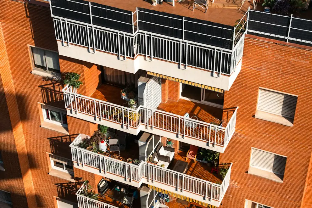 A red brick apartment building with stacked balconies containing potted plants and furniture